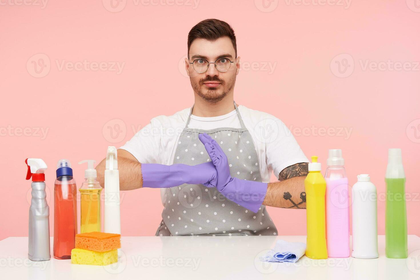 Indoor photo of agitated young bearded brunette man with short haircut looking menacingly at camera while sitting over pink background in rubber gloves