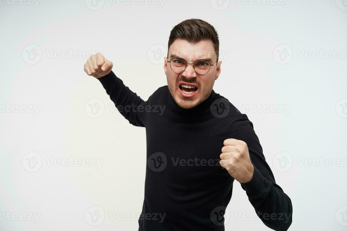 enojado joven bonito barbado morena chico con corto Corte de pelo ceñudo ferozmente su cara y levantamiento puños mientras siendo Listo a luchar, aislado terminado blanco antecedentes foto