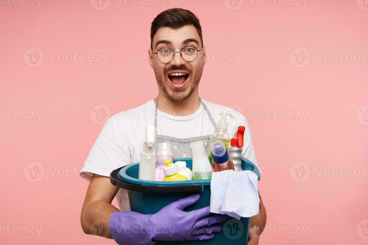 Overjoyed young handsome short brunette male in glasses dressed in apron and white t-shirt keeping busket with household chemicals and looking excitedly at camera with eyes and mouth opened photo