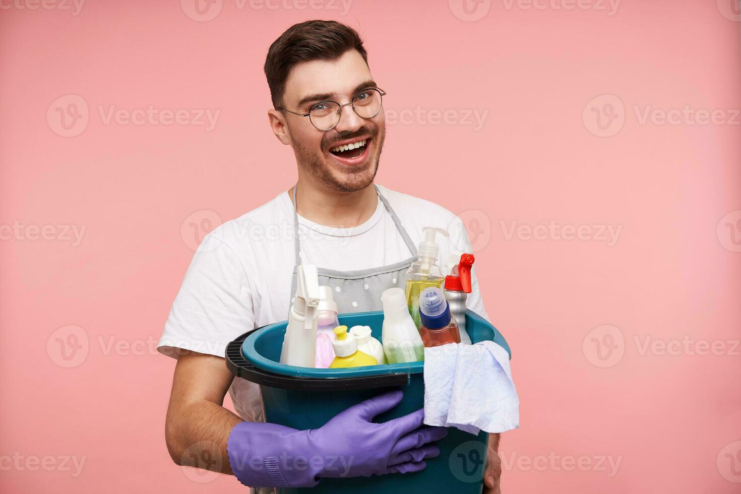 Indoor shot of glad young unshaved brunette man in uniform preparing for spring cleaning and holding household chemicals while posing over pink background photo