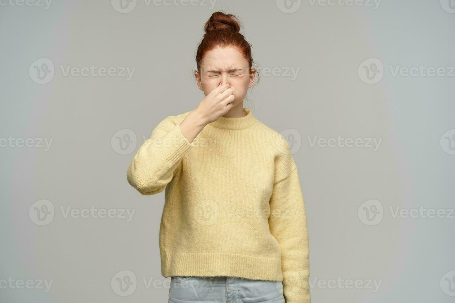 Portrait of sneezing, adult girl with ginger hair bun. Wearing yellow sweater. Squint her closed eyes, keep her nose closed and puffing cheeks. Holding breath. Stand isolated over grey background photo