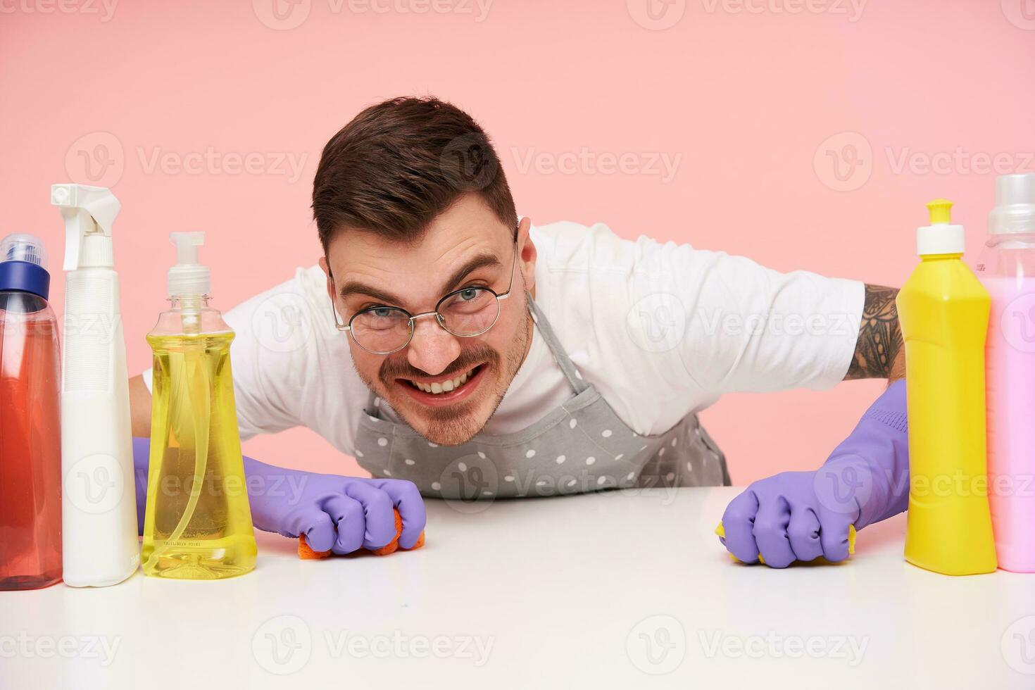 Portrait of young cheerful unshaved brunette guy with short haircut washing table and looking at camera with broad smile, sitting over pink background photo