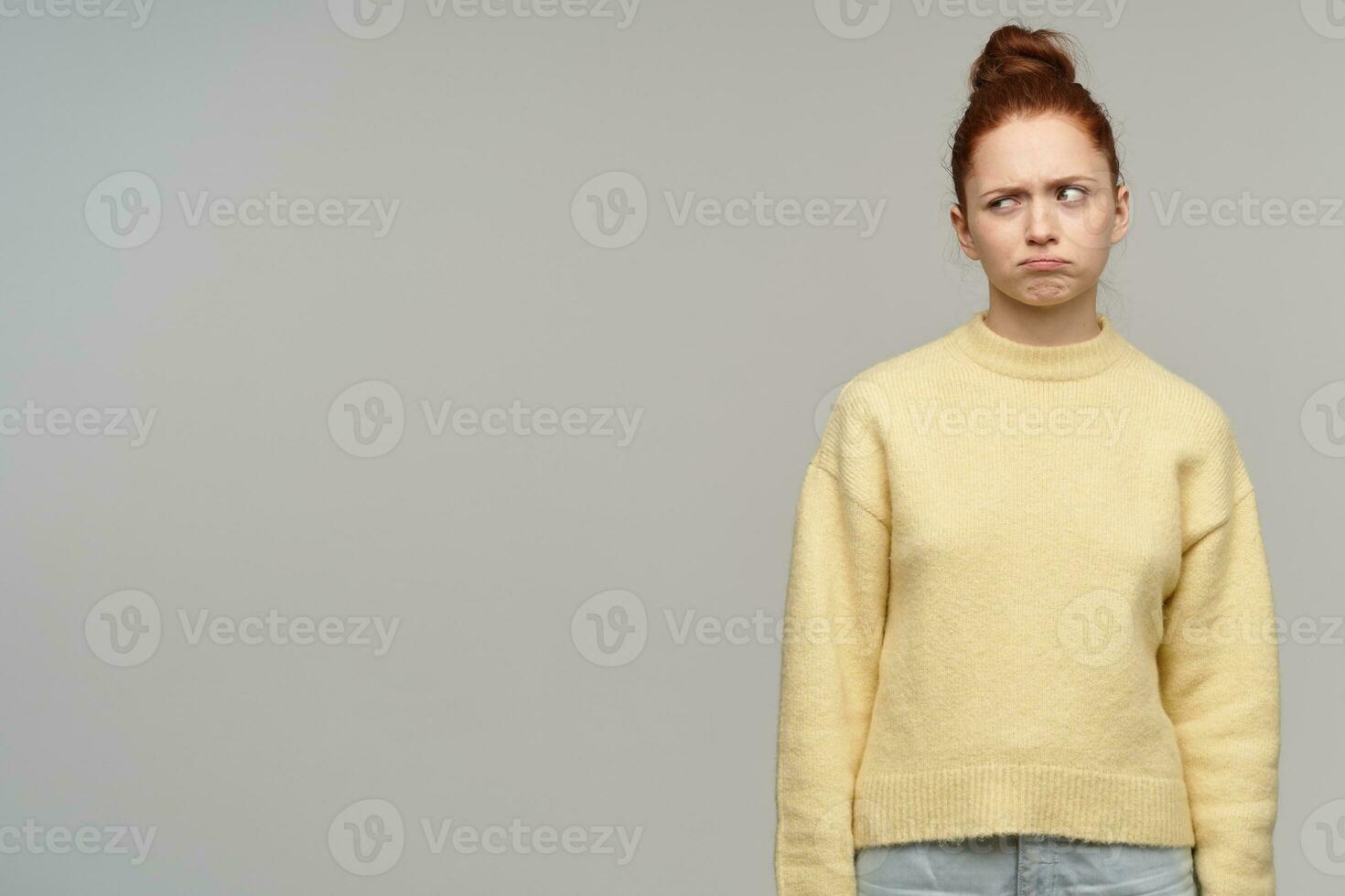 Unhappy lady, frowning upset woman with ginger hair bun. Wearing yellow sweater. Emotion concept. Watching to the left at copy space with wry face, isolated over grey background photo