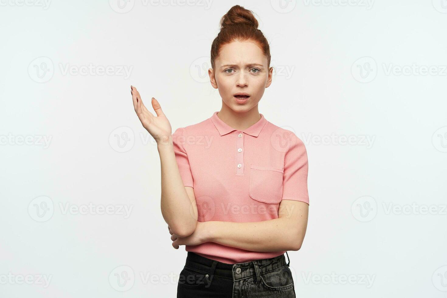 Unhappy lady, frowning woman with ginger hair bun. Wearing pink t-shirt and black jeans. Raise her hand in disagreement. Watching at the camera, isolated over white background. photo