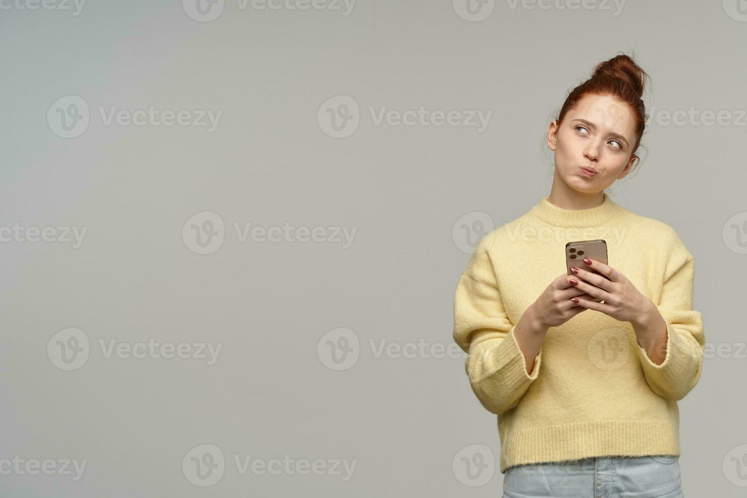 Portrait of attractive, cute girl with ginger hair bun. Wearing yellow sweater and holding a smartphone. Tilt her head and purse lips. Watching to the left at copy space, isolated over grey background photo