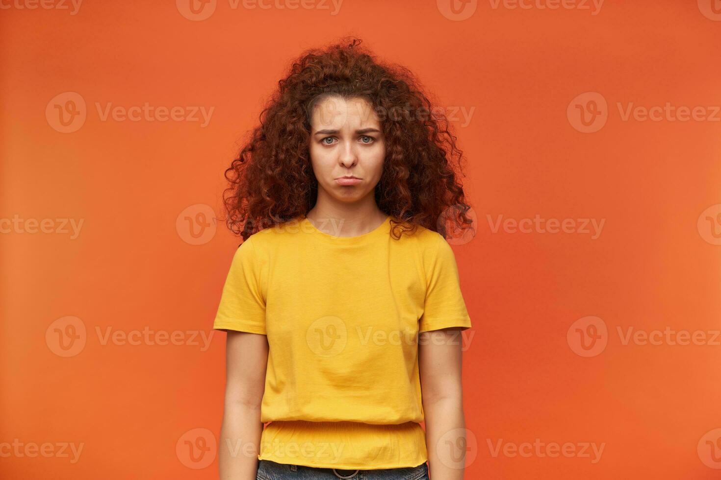 Unhappy looking woman, beautiful girl with curly ginger hair. Wearing yellow t-shirt. People and emotion concept. Pout her lip, upset. Watching at the camera isolated over orange background photo