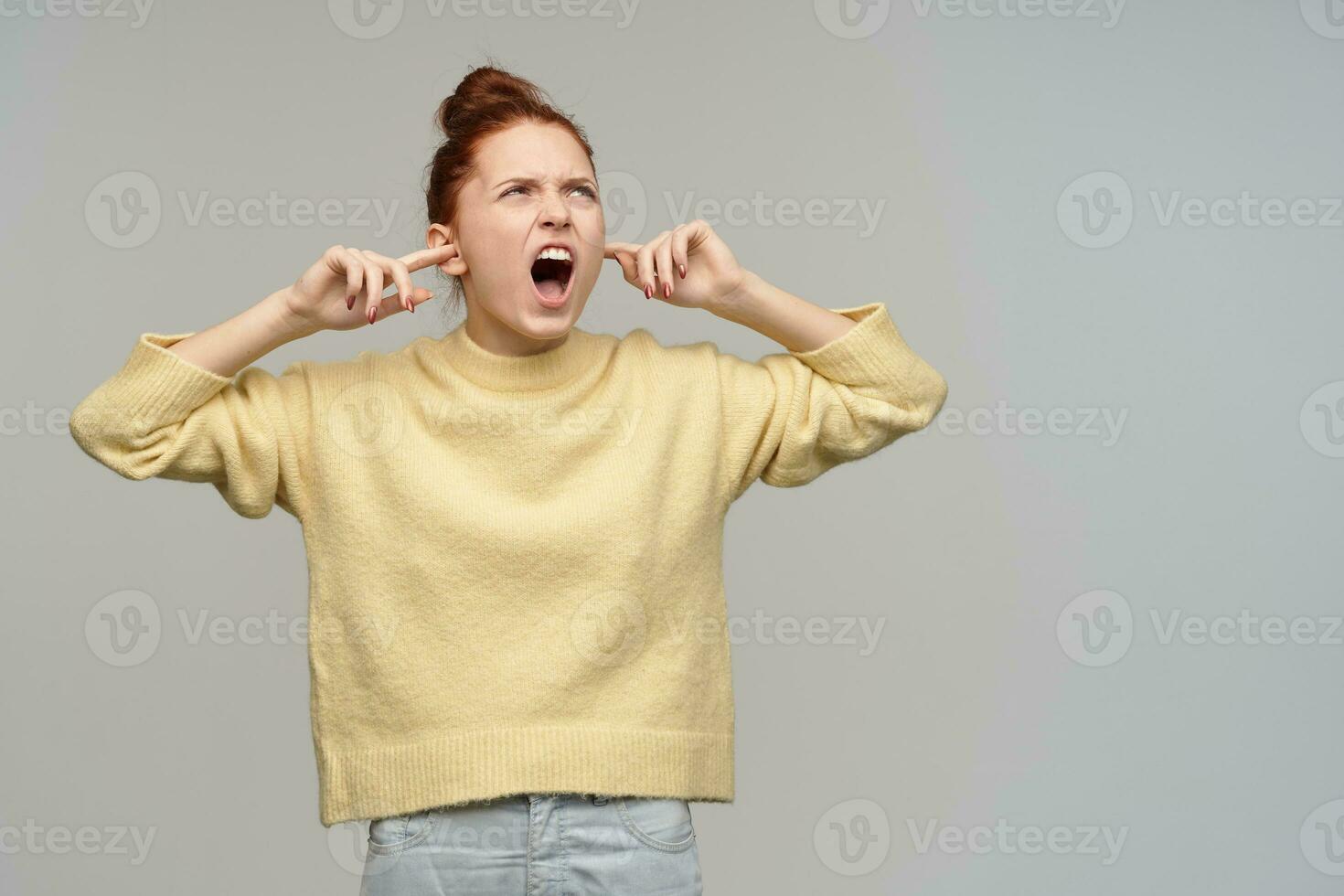 Unhappy, screaming girl with ginger hair gathered in a bun. Wearing pastel yellow sweater and jeans. Close her ears and yells. Watching to the right at copy space, isolated over grey background photo