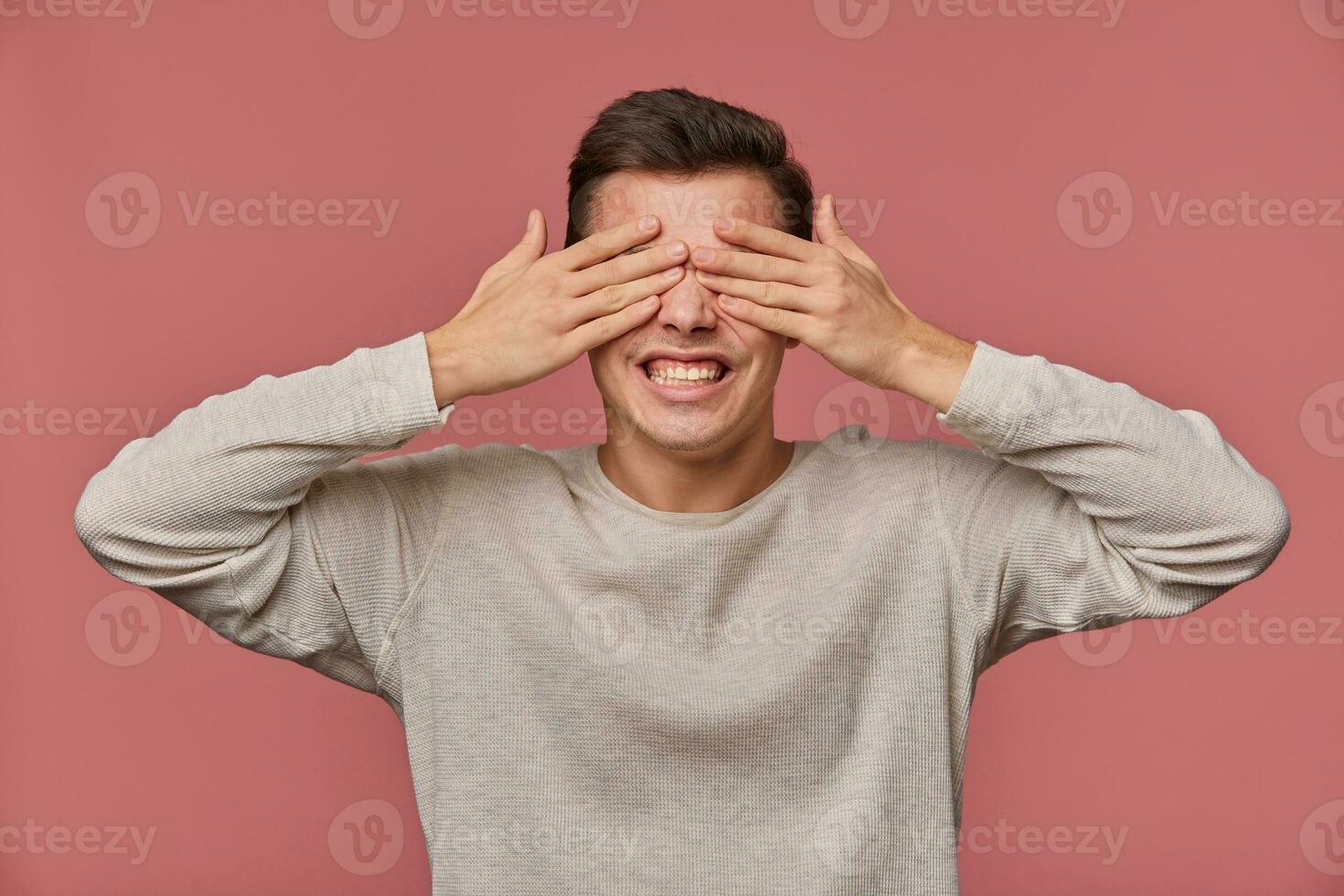 Photo of young surprised guy in blank long sleeve covers eyes with palms, stands over pink background with wide open eyes and screaming.