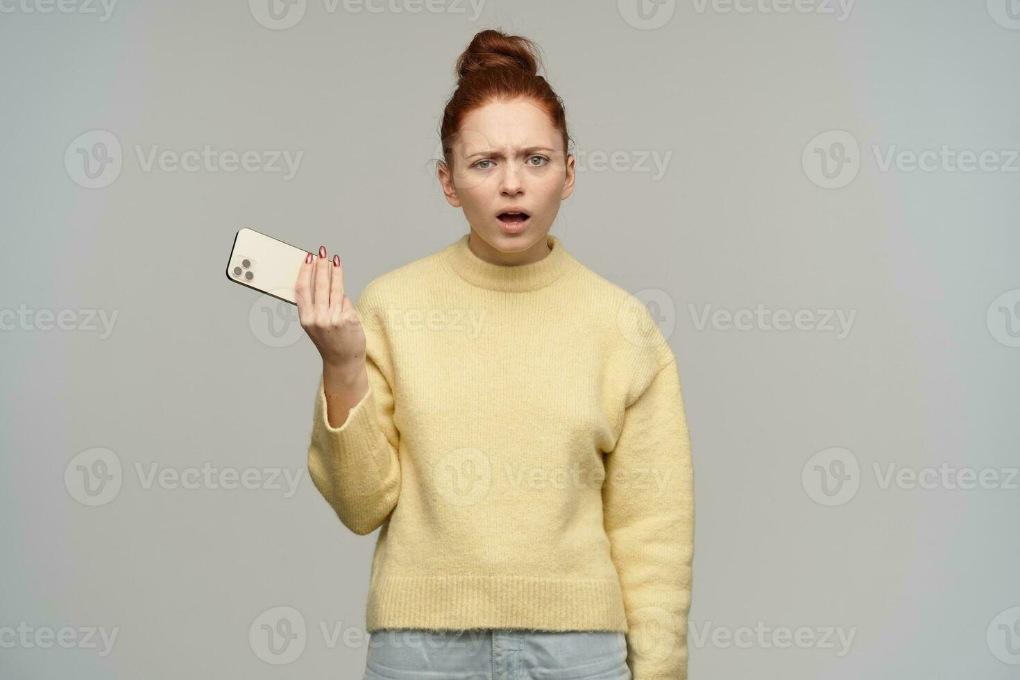 Confused lady, disconnected woman with ginger hair bun. Wearing yellow sweater and holding a smartphone. Frowning her face, bothered. Watching at the camera, isolated over grey background photo