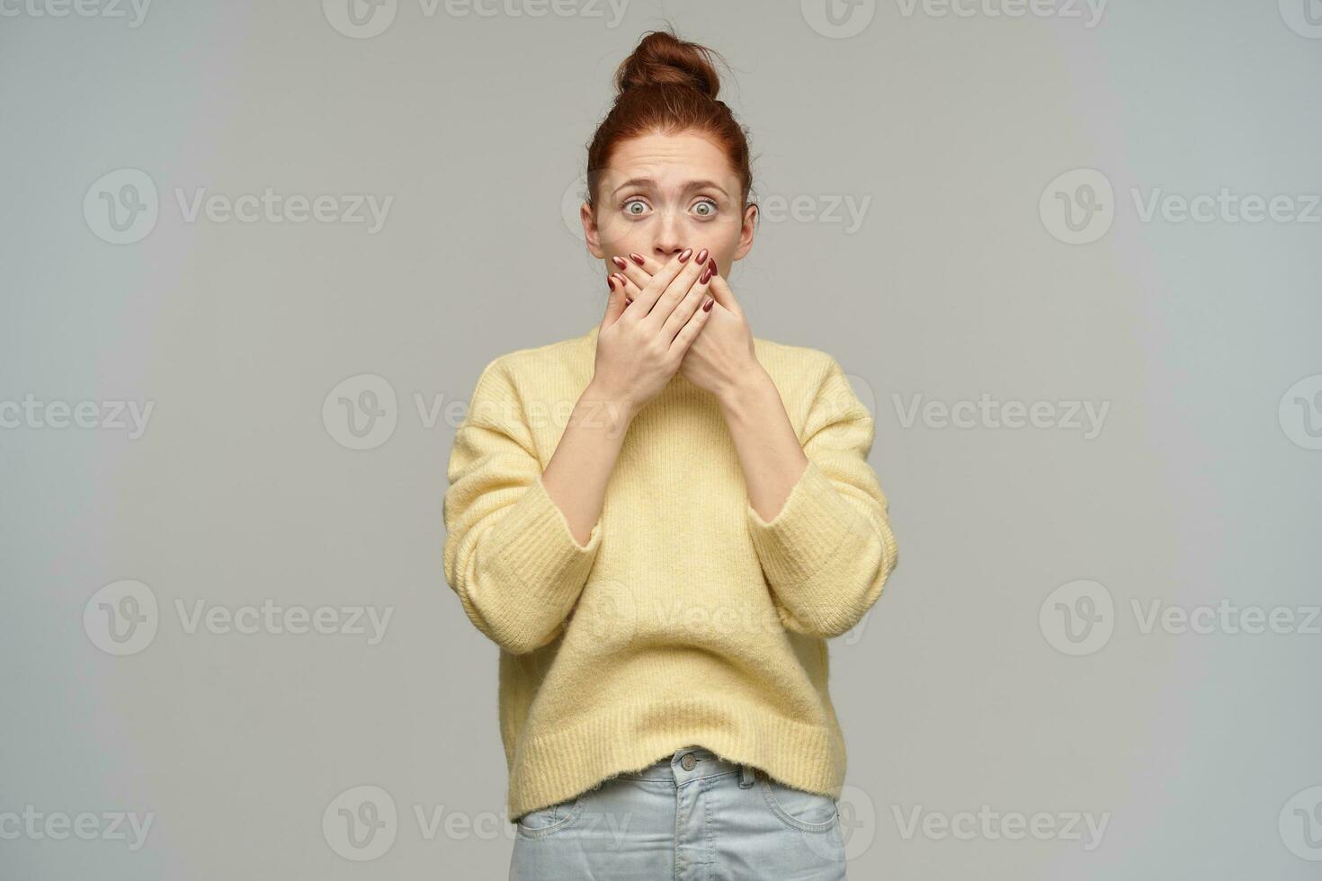 Shocked looking woman, beautiful girl with ginger hair gathered in a bun. Wearing pastel yellow sweater and jeans. Cover her mouth with palms. Watching at the camera isolated over grey background photo