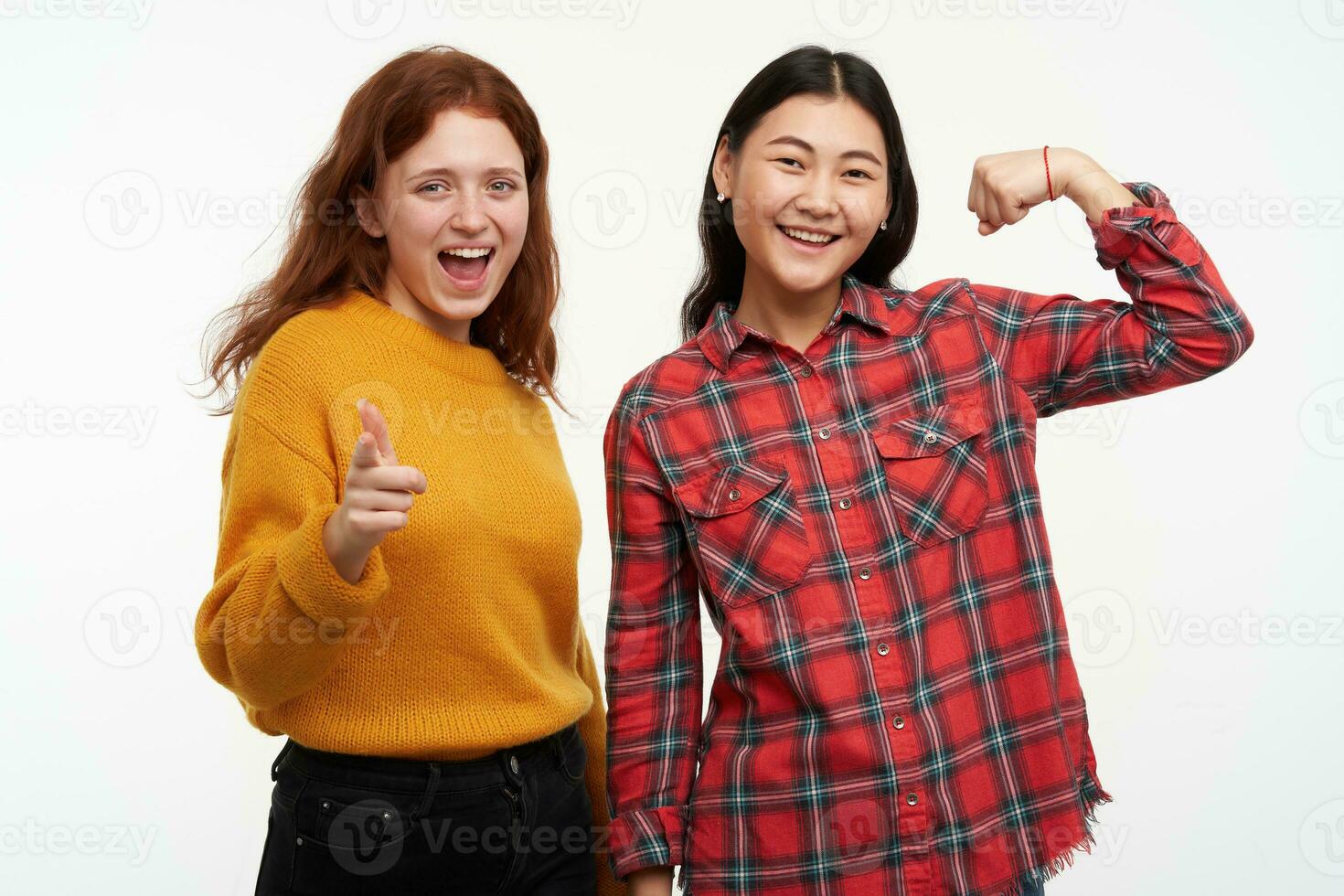 People and lifestyle concept. Two happy girls wearing yellow sweater and checkered shirt. Pointing at you with smile and friend shows biceps. Watching at the camera, isolated over white background photo