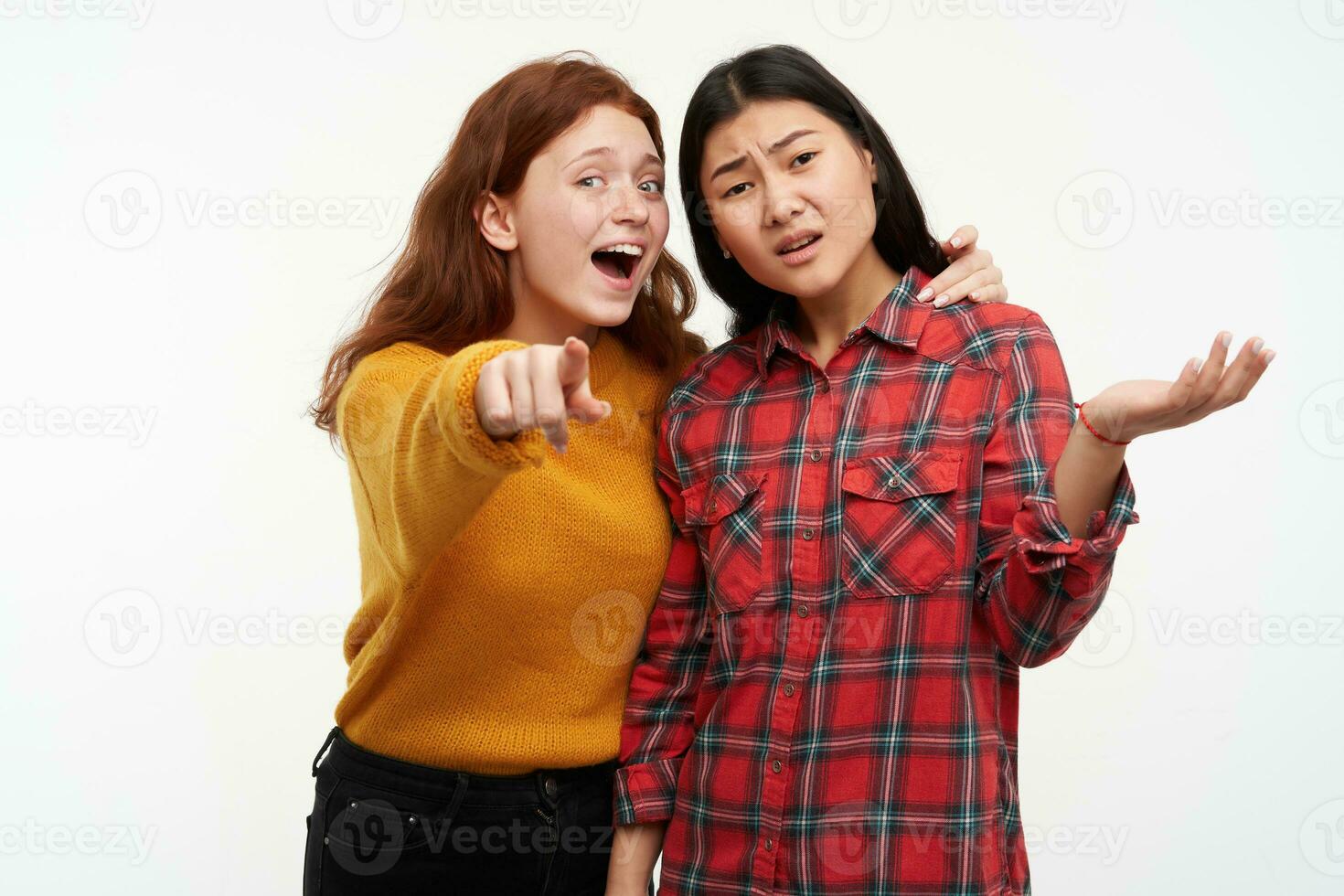 Two young women friends. Girl showing to her friend something, but another one can not see. Wearing yellow sweater and checkered shirt. Watching at the camera, isolated over white background photo