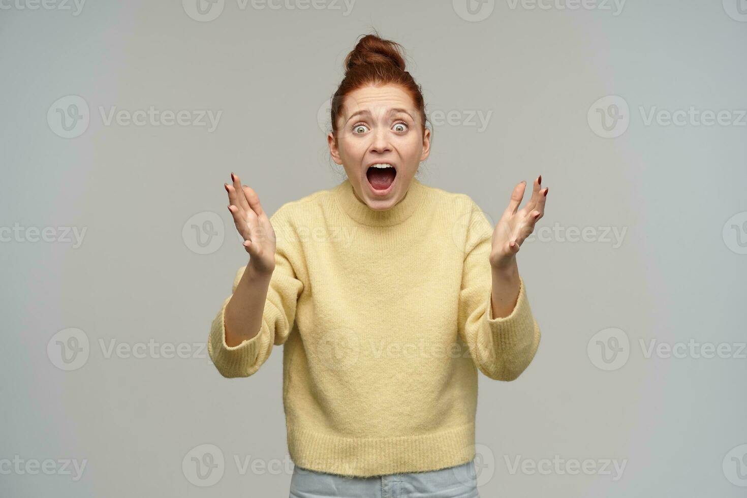 Horrified looking woman with ginger hair gathered in a bun. Wearing pastel yellow sweater and jeans. Screams in fear, terrified by what she see. Watching at the camera isolated over grey background photo