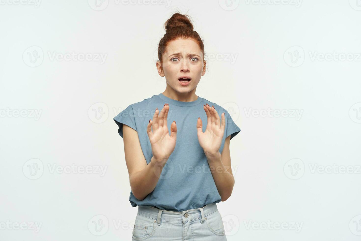 Afraid looking woman, beautiful girl with ginger hair gathered in a bun. Wearing blue t-shirt and jeans. Protect herself with hands. Watching horrified at the camera isolated over white background photo