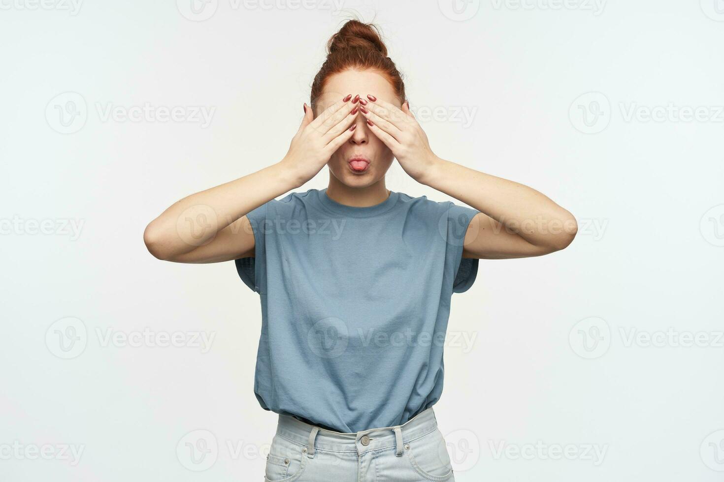 Portrait of attractive, playful redhead girl with hair gathered in a bun. Wearing blue t-shirt and jeans. Cover her eyes with palms and showing a tongue. Stand isolated over white background photo