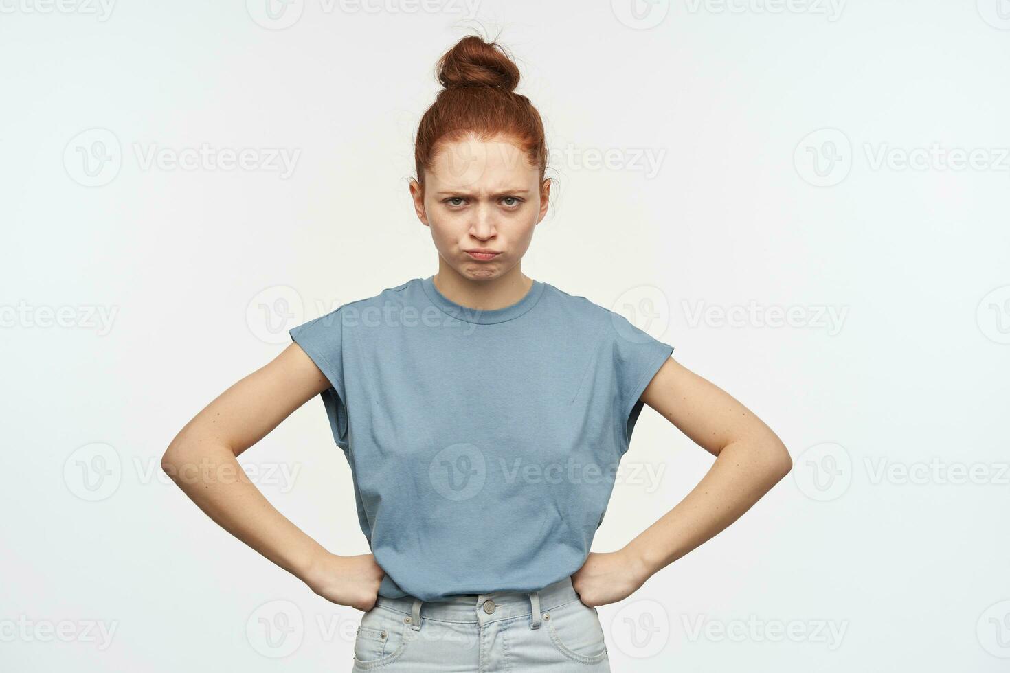 Portrait of upset, adult redhead girl with hair gathered in a bun. Wearing blue t-shirt and jeans. Put hands on her hips and frowning. Watching at the camera isolated over white background photo