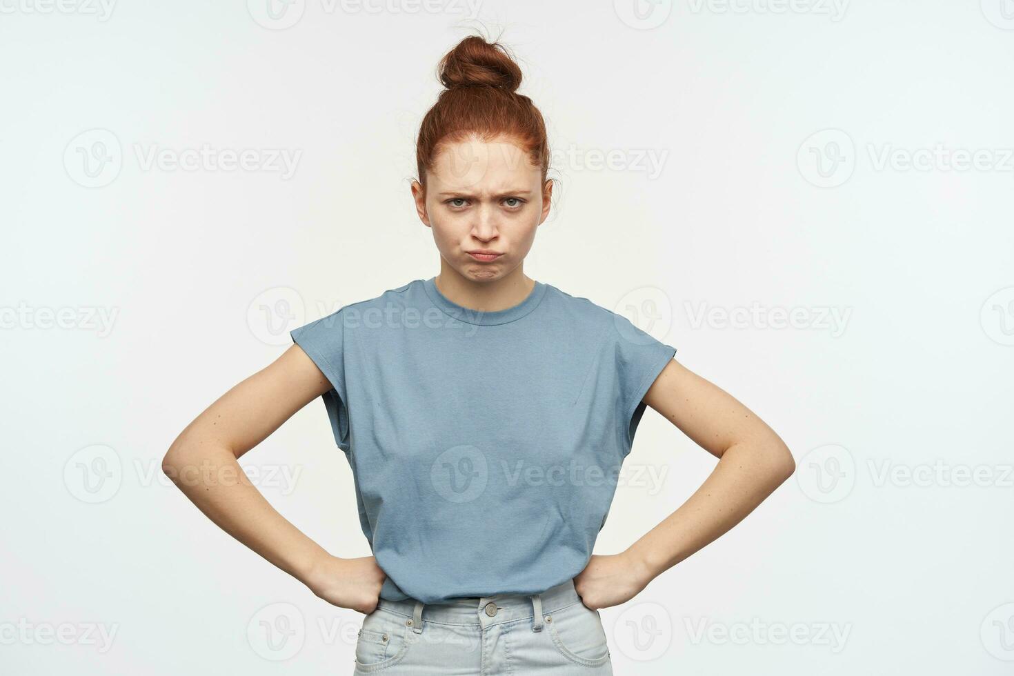 retrato de decepcionado, adulto pelirrojo niña con pelo reunido en un bollo. vistiendo azul camiseta y vaqueros. poner manos en su caderas y ceñudo. acecho a el cámara aislado terminado blanco antecedentes foto