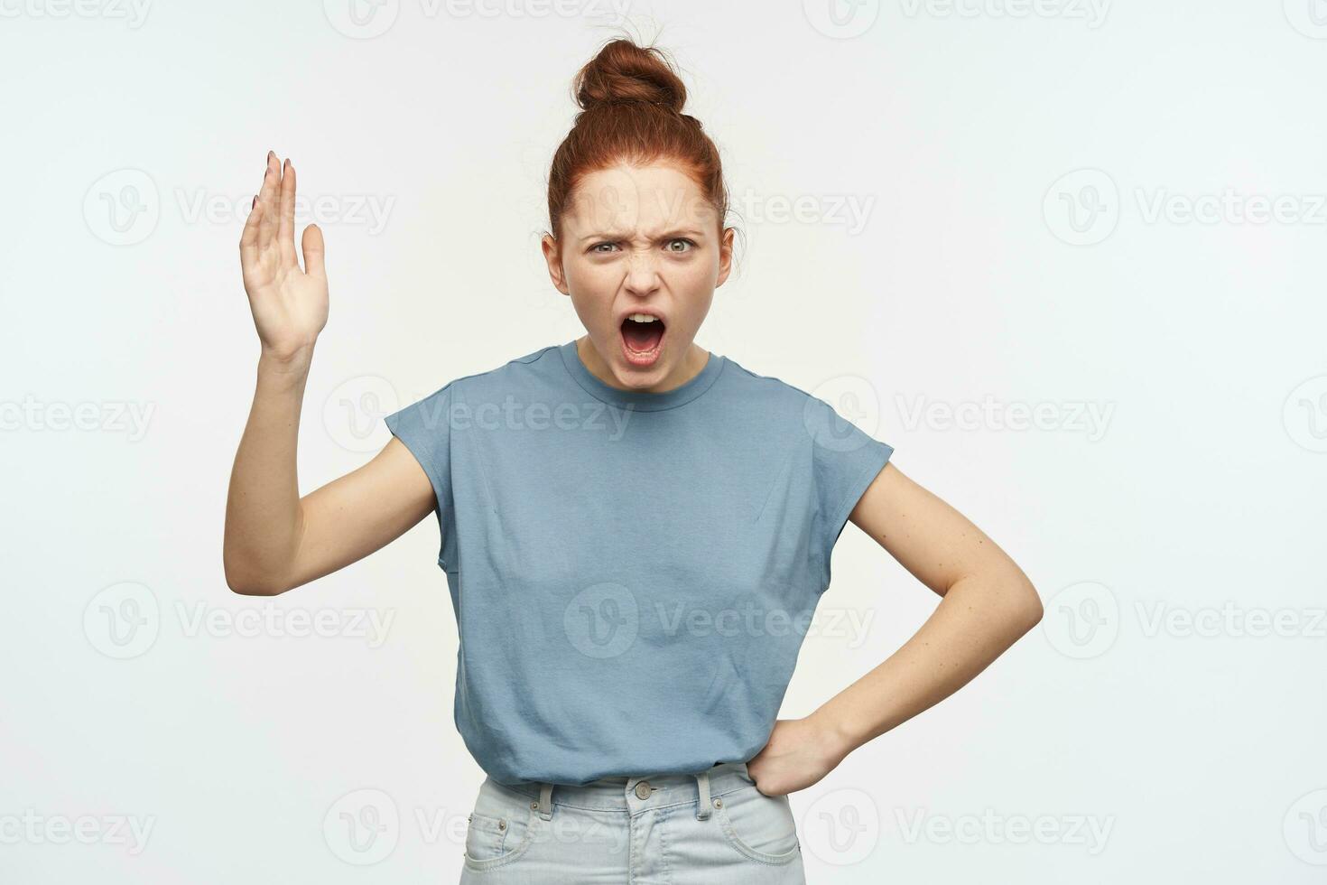 Teenage, angry looking woman with ginger hair gathered in a bun. Wearing blue t-shirt and jeans. Raise her hand and put another on a hip, shout. Watching at the camera isolated over white background photo