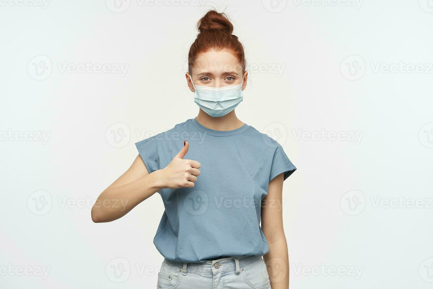 Nice looking woman, happy girl with ginger hair gathered in a bun. Wearing blue t-shirt, jeans and protective face mask. Showing thumb up. Watching at the camera isolated over white background photo