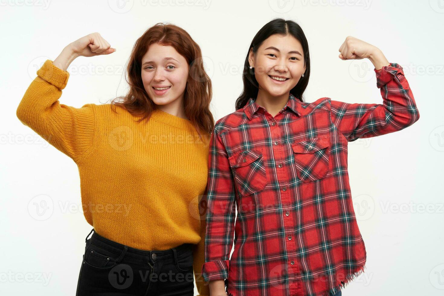 Two young, happy women friends. Wearing yellow sweater and checkered shirt. Showing how strong they are, muscles. Full of energy. People concept. Watching at the camera, isolated over white background photo
