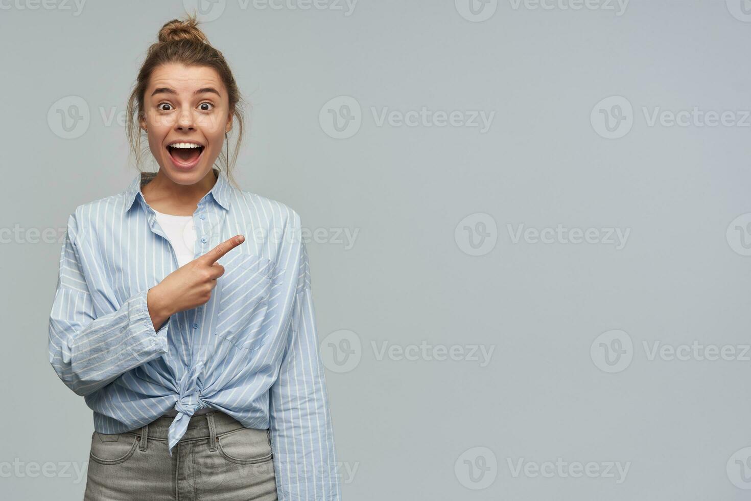 Cheerful and happy woman with blond hair gathered in bun. Wearing striped knotted shirt. Pointing with index finger to the right at copy space. Watching at the camera, isolated over grey background photo