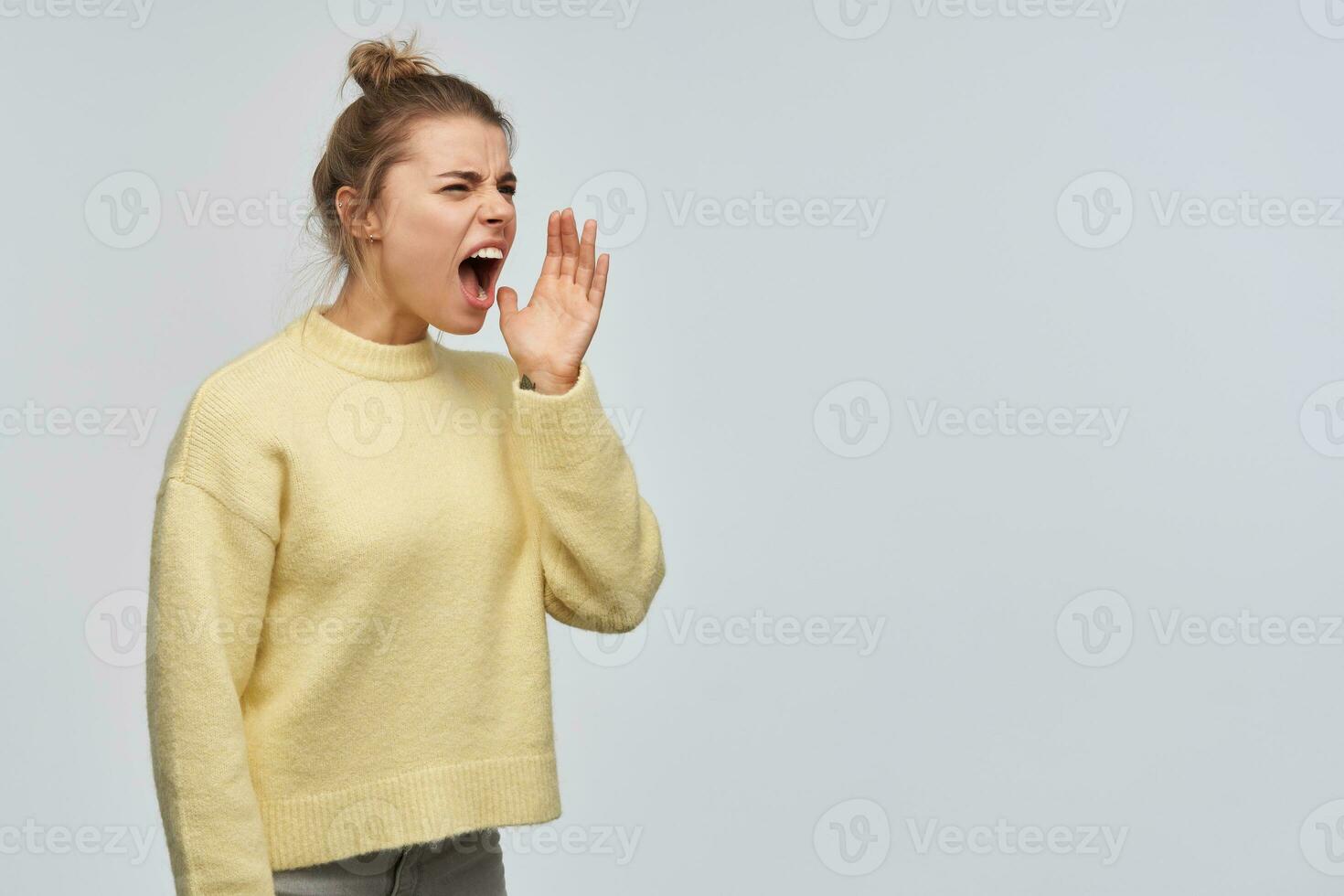 Portrait of angry, adult girl with blond hair gathered in bun. Wearing yellow sweater. Holds palm next to her mouth and shout to the right at copy space. Stand isolated over white background photo