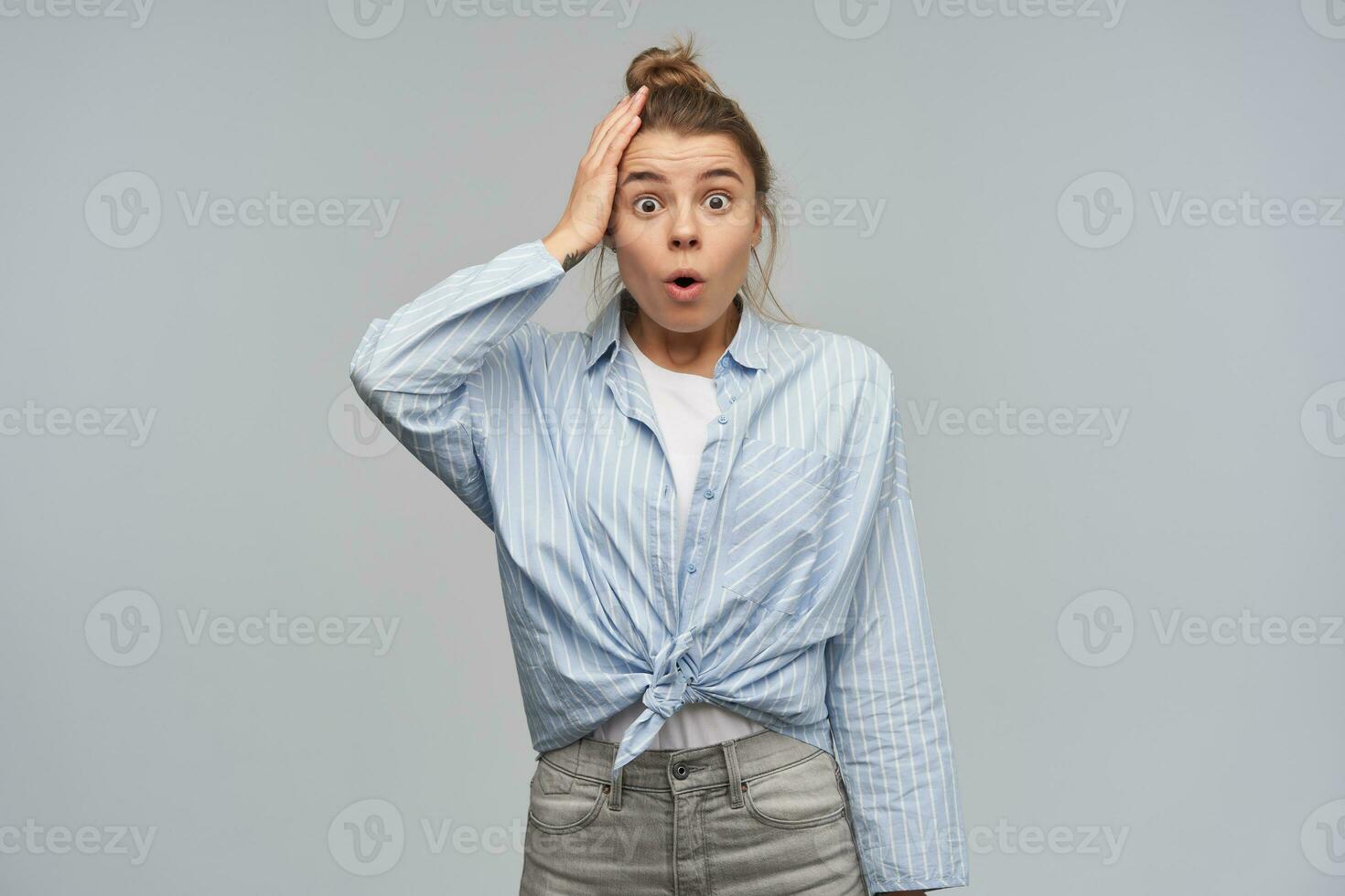 Teenage girl, shocked looking woman with blond hair gathered in bun. Wearing striped knotted shirt. Touching her head. Forget to turn off a stove. Watching at the camera, isolated over grey background photo