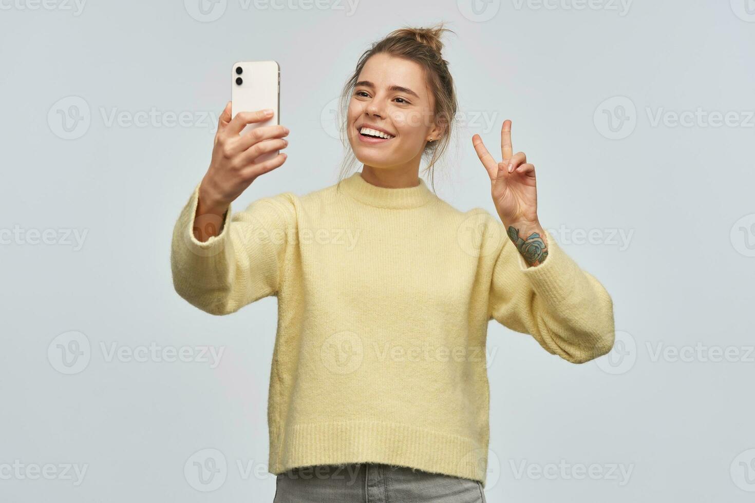 Portrait of attractive, adult girl with blond hair gathered in bun and tattoo. Wearing yellow sweater and holding a smartphone. Making selfie. Showing peace sign. Stand isolated over white background photo