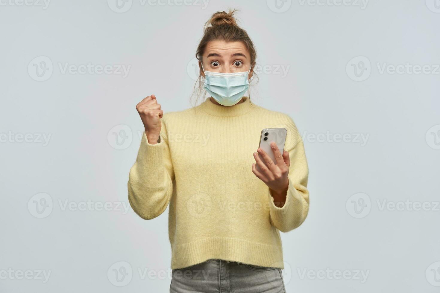 Excited, happy woman with blond hair gathered in bun. Wearing yellow sweater and protective face mask. Holding a smartphone and clench her fist. Watching at the camera, isolated over white background photo