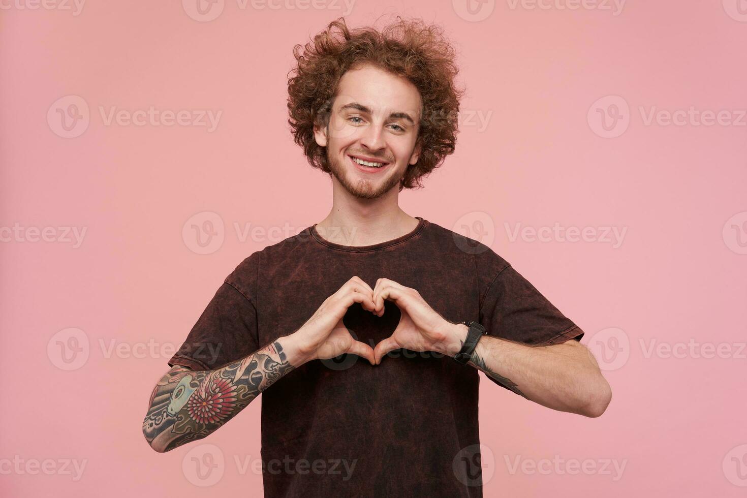 Young, happy man with brunette curly hair, beard and tattoos. Wearing dark red t-shirt. Emotion concept. Showing love sign with fingers. Watching at the camera isolated over pastel pink background photo