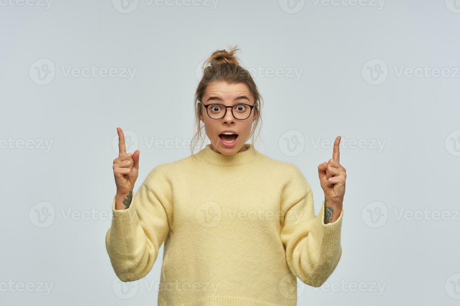 Teenage, shocked girl with blond hair gathered in bun and tattoo. Wearing yellow sweater and eyewear. Pointing index fingers up at copy space. Watching at the camera, isolated over white background photo