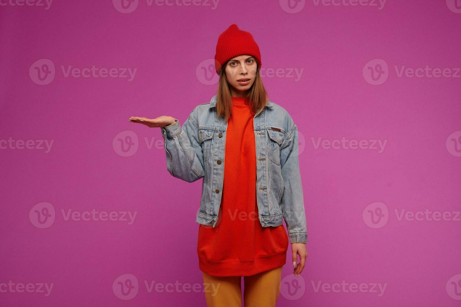 Nice looking beautiful girl with brunette hair. Wearing jeans jacket, yellow pants, red sweater and hat. Pretend to hold something on a palm. Watching at the camera isolated over purple background photo