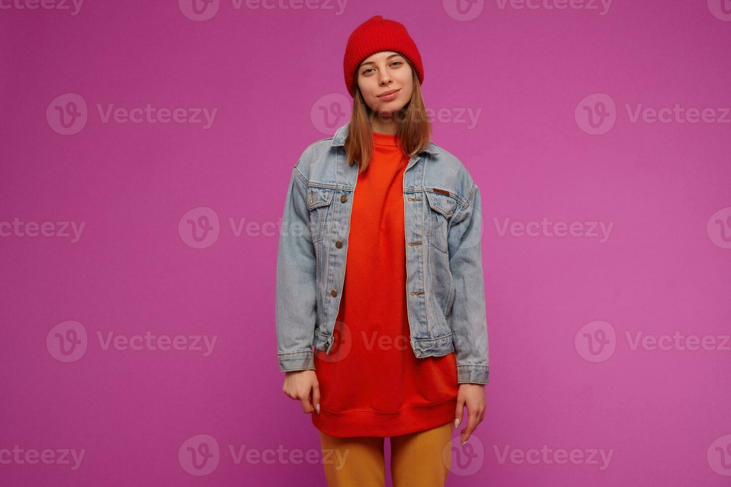 Teenage girl, happy looking woman with brunette long hair. Wearing jeans jacket, yellow pants, red sweater and hat. Watching with confidence at the camera isolated over purple background photo