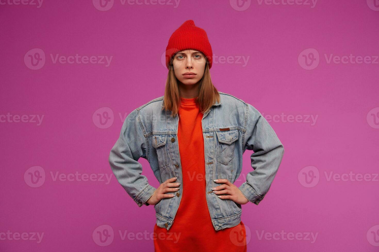 Portrait of young woman with brunette long hair. Wearing jeans jacket, red sweater and hat. Holds hands on a waist, frown. Watching suspiciously at the camera isolated over purple background photo