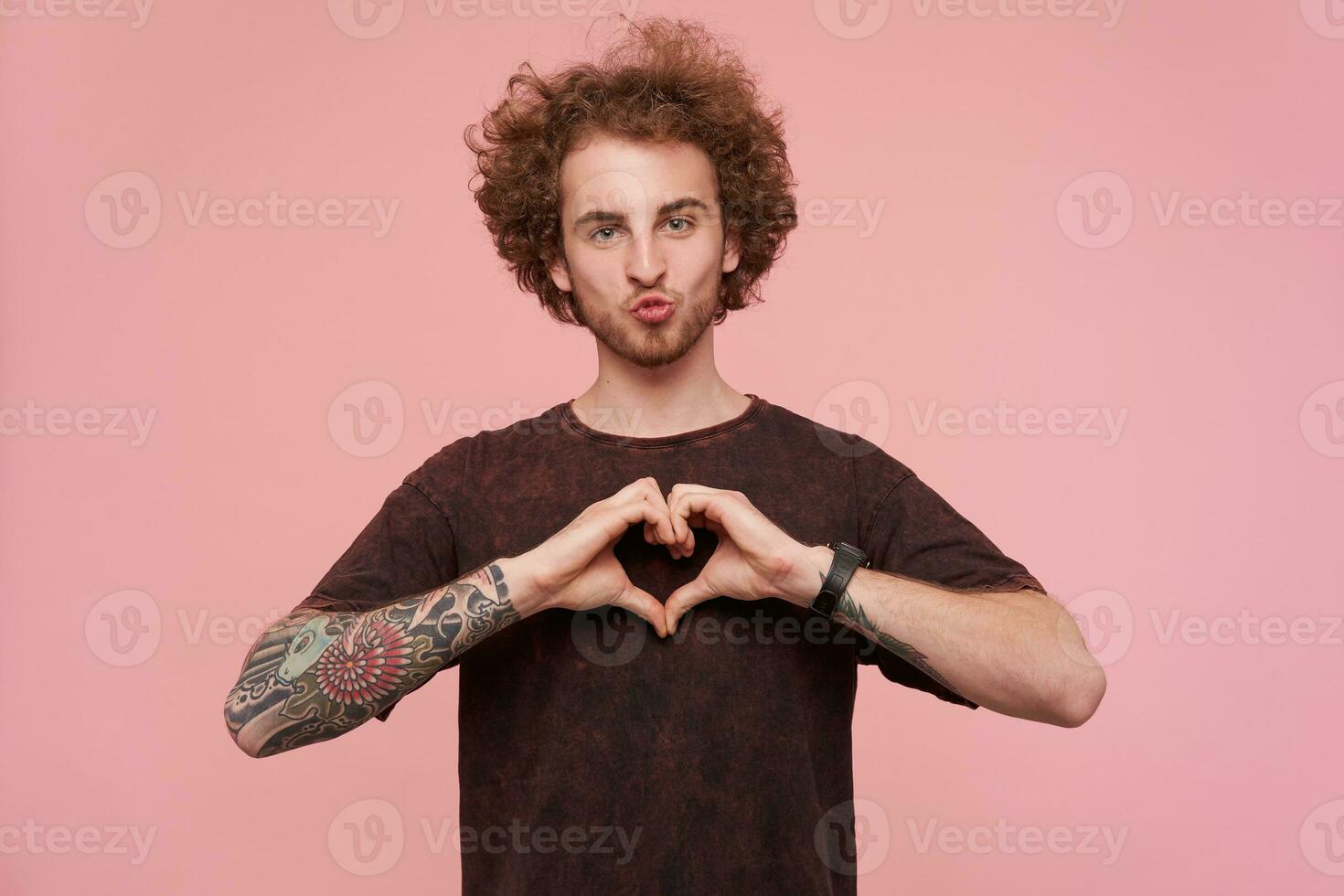 Handsome guy with tattoos, brunette curly hair and beard. Wearing dark red t-shirt. Showing love sign with fingers and try to kiss. Watching at the camera isolated over pastel pink background photo