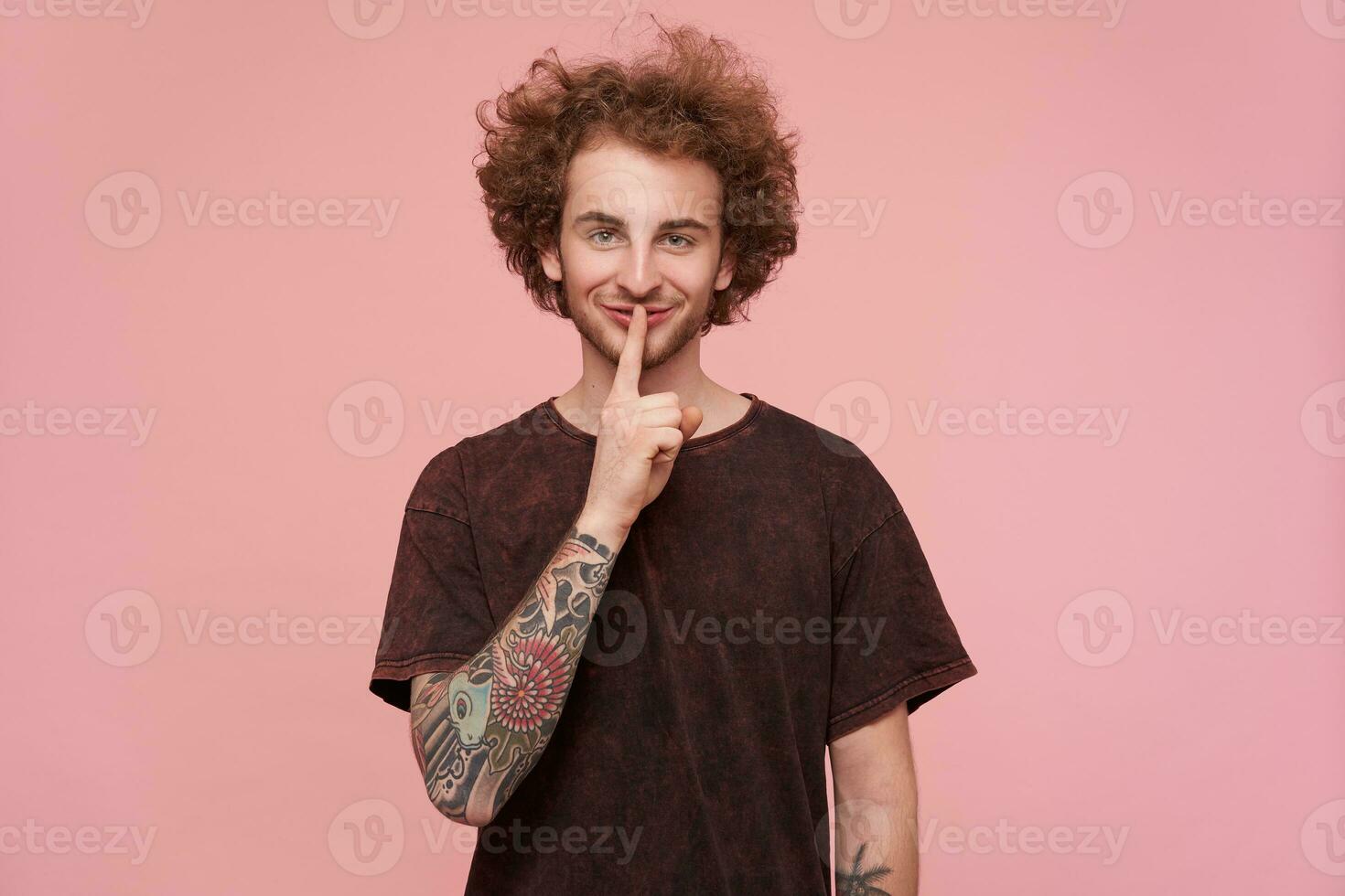 Young man, cool guy with brunette curly hair, beard and tattoos. Wearing dark red t-shirt. Emotion concept. Showing silence sign and smile. Watching at the camera isolated over pastel pink background photo