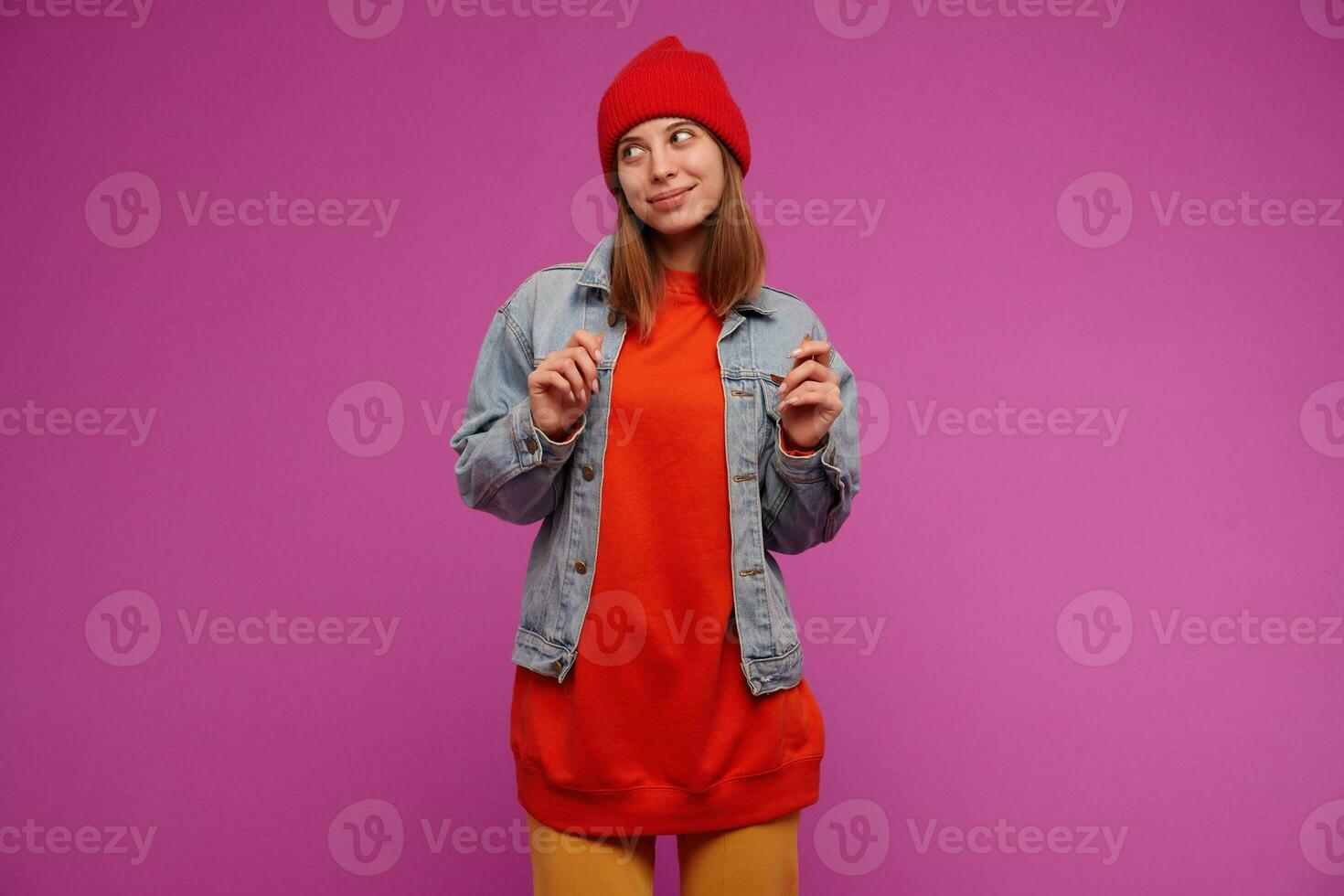Teenage girl, happy looking woman with brunette long hair. Wearing jeans jacket, yellow pants, red sweater and hat. Watching to the left at copy space, isolated over purple background photo