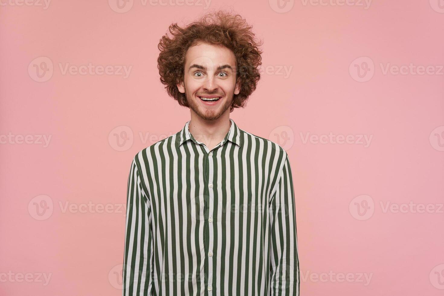 Young cool guy with brunette curly hair and beard. Wearing shirt with black and white stripes. Emotion and people concept. Excited, smile. Watching at the camera isolated over pastel pink background photo