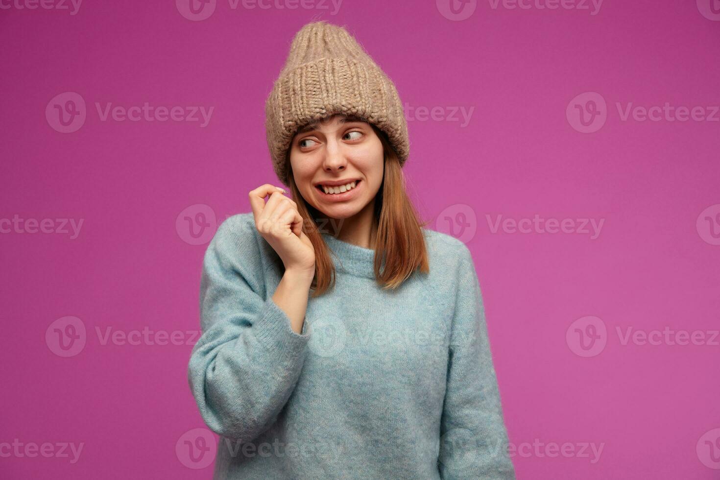 Portrait of attractive woman with brunette long hair. Wearing blue sweater and knitted hat. Emotion concept. Shrugs her shoulders. Watching to the left at copy space, isolated over purple background photo