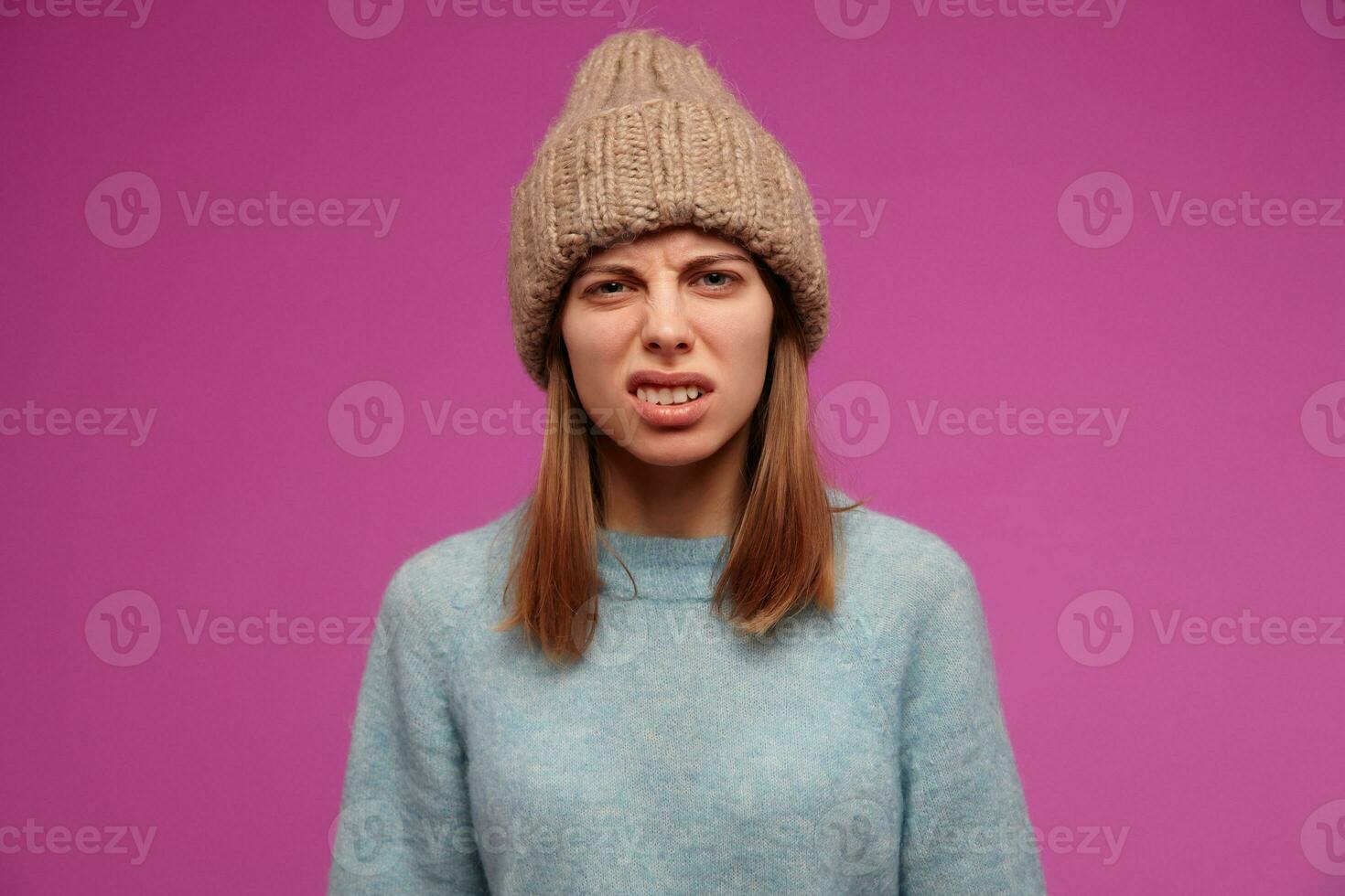 Angry looking woman, frowning girl with brunette hair. Wearing blue sweater and knitted hat. Emotion concept. Lifts her lip in disgust. Watching at the camera isolated over purple background photo