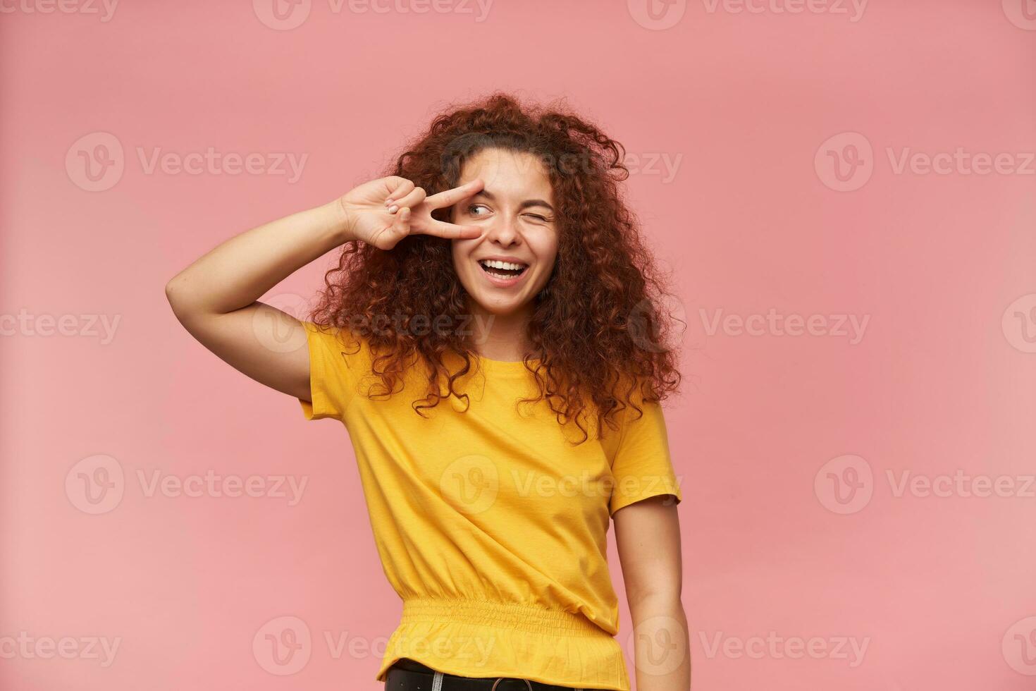 Portrait of attractive, redhead girl with curly hair. Wearing yellow t-shirt. Emotion concept. Showing peace sign over her eye. Watching to the left at copy space, isolated over patel pink background photo