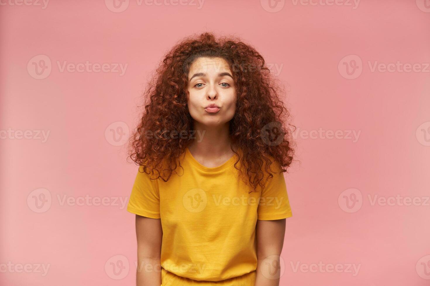 Portrait of attractive, redhead girl with curly hair. Wearing yellow t-shirt. People and emotion concept. Wants to kiss you. Watching at the camera isolated over pastel pink background photo