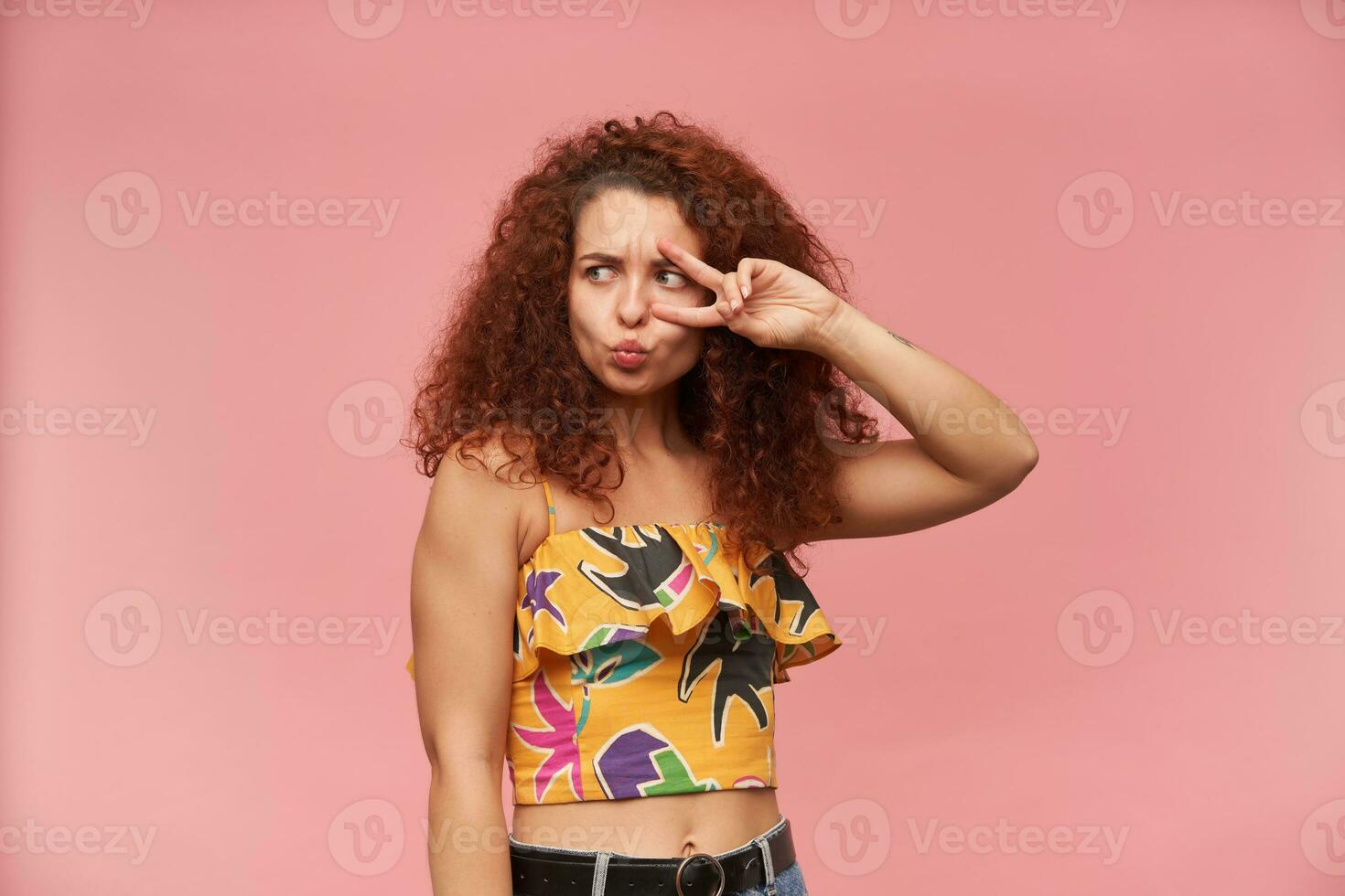 Cute girl with curly ginger hair. Wearing colorful off-shoulder blouse. Showing peace sign over eye. Fold lips for a kiss. Watching to the left at copy space, isolated over patel pink background photo