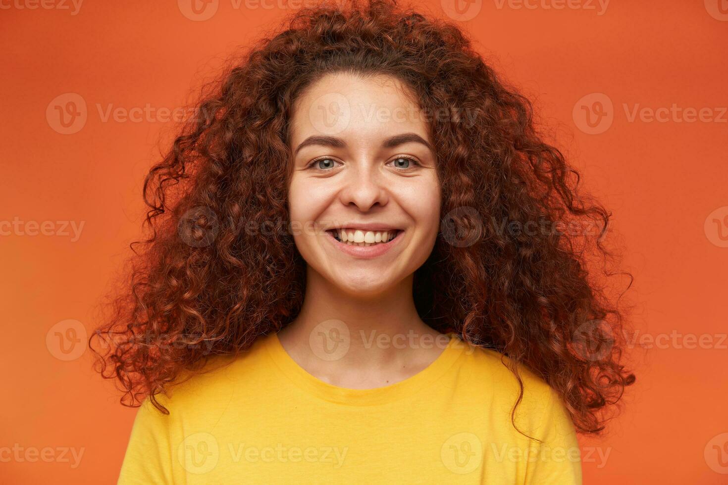 Teenage girl, happy looking woman with ginger curly hair. Wearing yellow t-shirt. People and emotion concept. Watching at the camera with toothy smile, close up, isolated over orange background photo