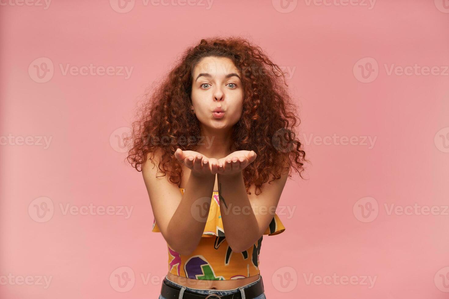 Portrait of lovely, redhead girl with curly hair. Wearing colorful off-shoulder blouse. Emotion concept. Sending air kiss, blow over palms. Watching at the camera isolated over pastel pink background photo