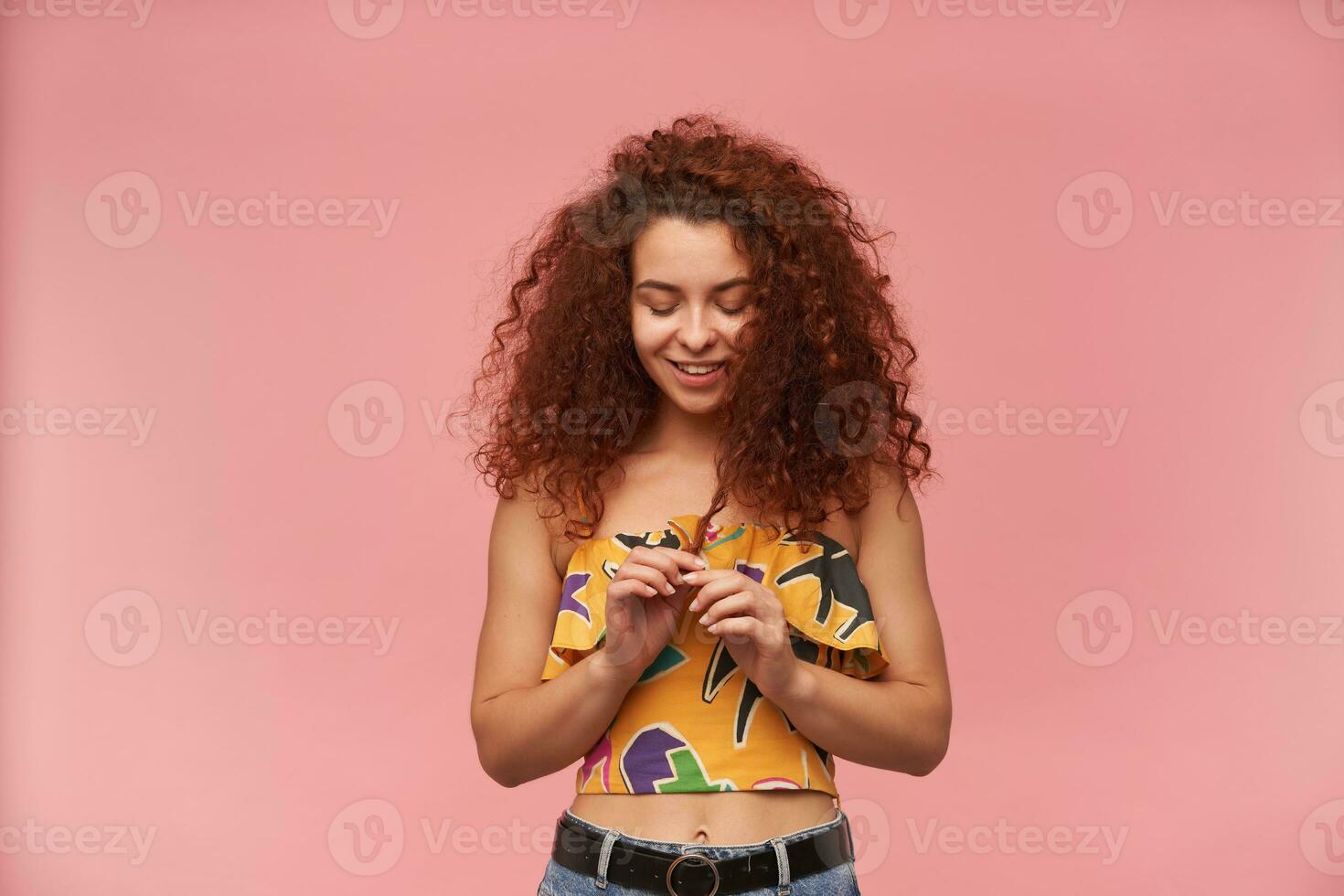 Portrait of attractive, redhead girl with curly hair. Wearing colorful off-shoulder blouse. People and emotion concept. Shyly playing with strand of hair. Stand isolated over pastel pink background photo