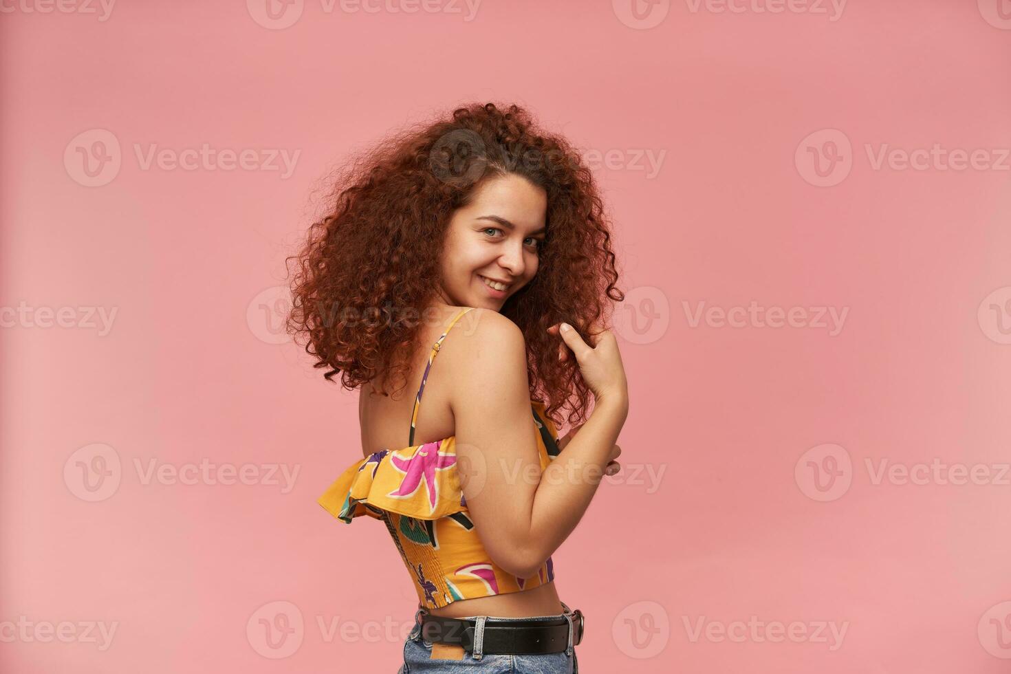 Shy looking woman, beautiful girl with curly ginger hair. Wearing colorful off-shoulder blouse. People and emotion concept. Watching at the camera over shoulder, isolated over pastel pink background photo