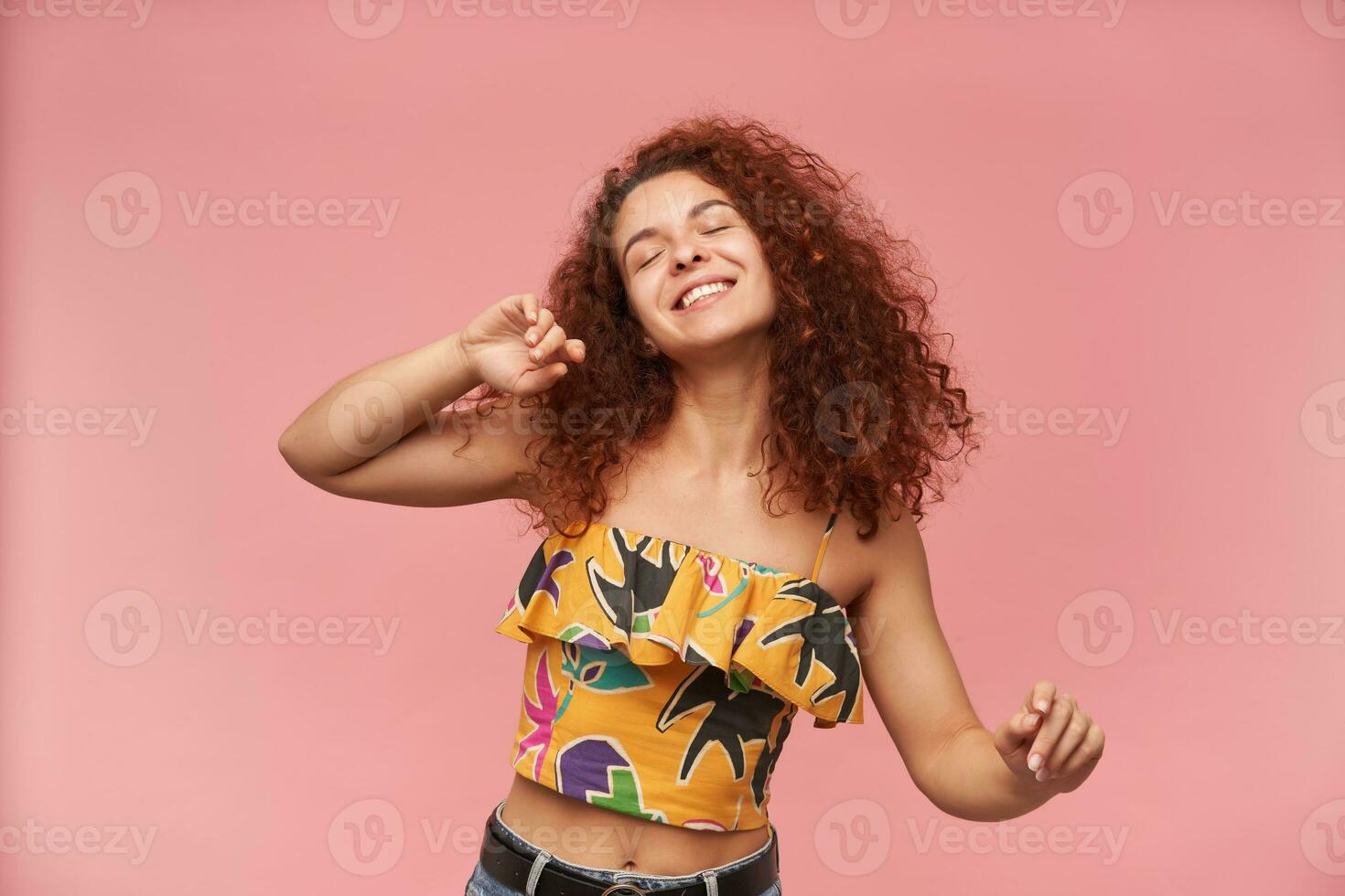 Portrait of attractive, redhead girl with curly hair. Wearing colorful off-shoulder blouse. People and emotion concept. Dancing with closed eyes. Stand isolated over pastel pink background photo