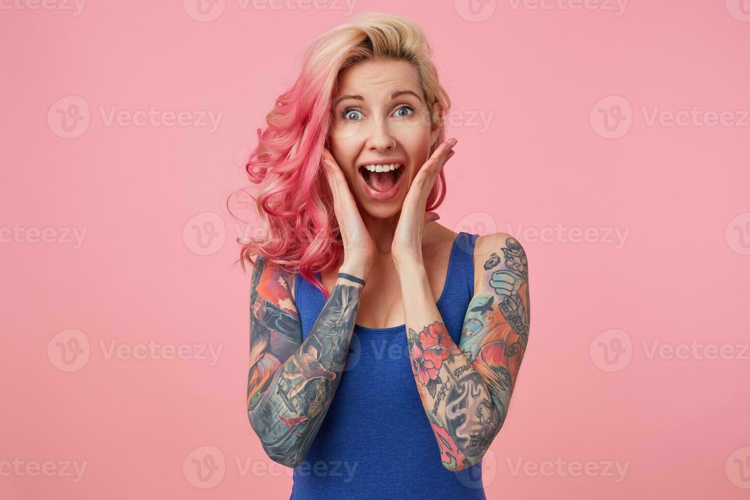 Portrait of young happy amazed nice pink haired lady, with palms on cheeks, her boyfriend proposed to her, looks at the camera with wide open mouth, feels happiness, stands over pink background. photo