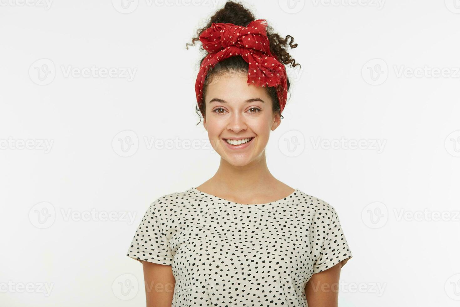 bonito encantador niña con marrón Rizado pelo reunido en un bollo agradablemente sonrisas, vestido en un blanco camiseta con negro polca puntos, en su cabeza un rojo bufanda con blanco polca puntos, terminado blanco antecedentes foto